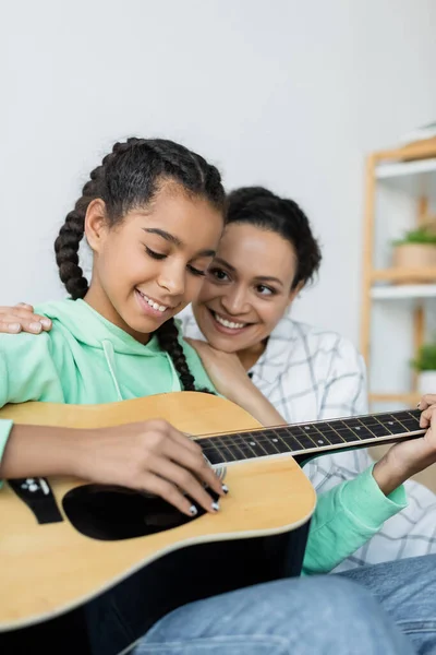 Afrikanisch-amerikanisches Teenager-Mädchen spielt Akustikgitarre in der Nähe einer glücklichen Mutter zu Hause — Stockfoto