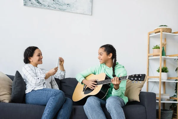African american woman with smartphone taking photo of daughter playing guitar at home — Stock Photo