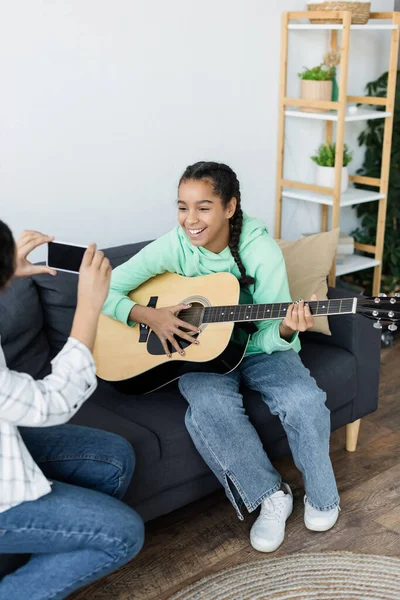 Femme prenant une photo d'une adolescente afro-américaine jouant de la guitare sur le canapé — Photo de stock