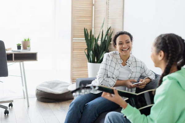 Blurred african american girl playing guitar near laughing mom sitting on couch with cellphone — Stock Photo