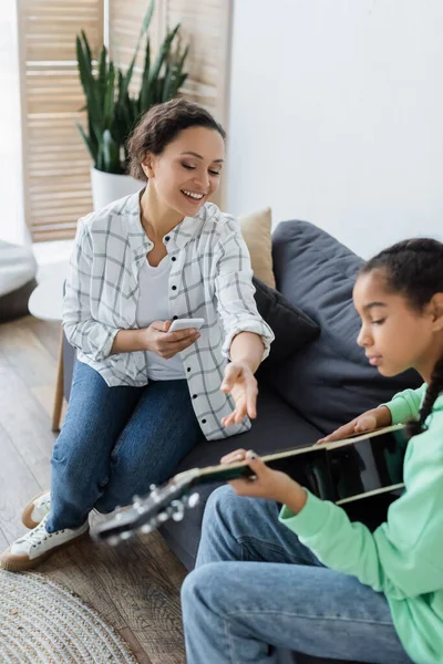 African american woman with smartphone pointing with hand near daughter playing acoustic guitar — Stock Photo