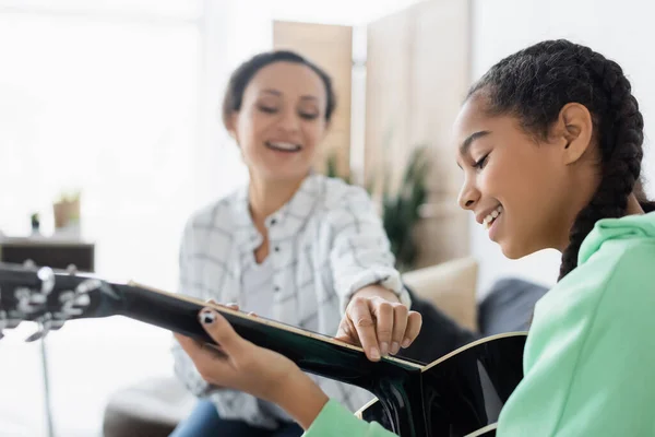 Blurred african american woman touching strings on guitar near smiling daughter — Stock Photo