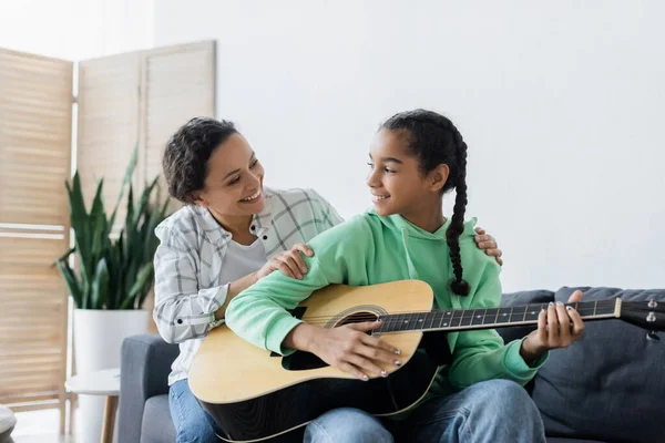 Sonriente africana americana chica mirando feliz mamá mientras jugando guitarra en casa - foto de stock