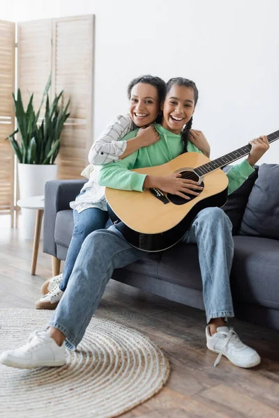 Erfreut afrikanisch-amerikanische Frau blickt in die Kamera, während sie Tochter umarmt, die auf dem Sofa Gitarre spielt — Stockfoto