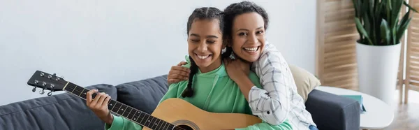 Mujer afroamericana emocionada mirando a la cámara mientras abraza a su hija tocando la guitarra acústica, pancarta - foto de stock