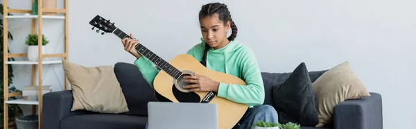 African american teen girl sitting on couch with pillows and learning to play guitar near laptop, banner — Stock Photo