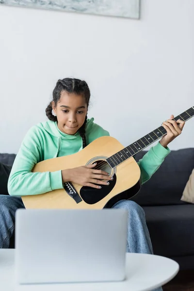 Focada menina afro-americana olhando para laptop borrado enquanto aprende a tocar guitarra em casa — Fotografia de Stock