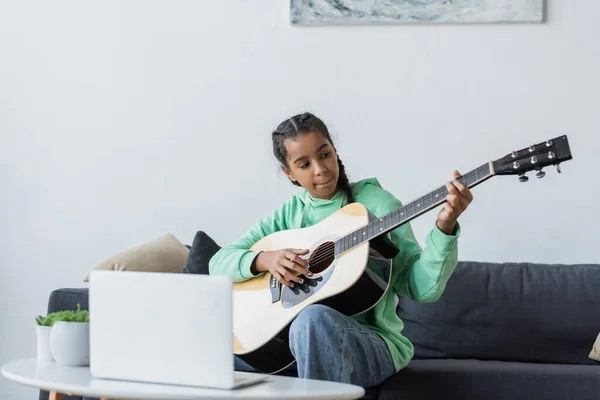 African american teenage girl sitting on couch near laptop and learning to play guitar — Stock Photo