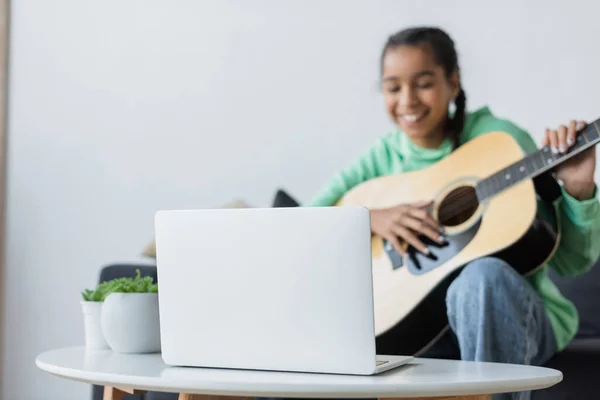 Borrosa afroamericana adolescente sonriendo mientras aprende a tocar la guitarra acústica cerca de la computadora portátil - foto de stock