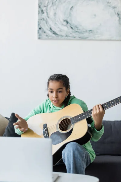 Afro-américaine adolescent fille pointant vers ordinateur portable tout en apprenant à jouer de la guitare à la maison — Photo de stock