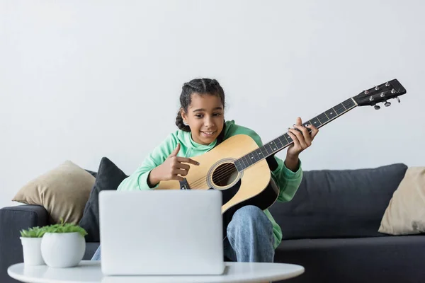 Lächelndes afrikanisch-amerikanisches Mädchen, das auf Laptop schaut und auf Gitarre zeigt, während es zu Hause spielen lernt — Stockfoto