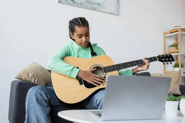 Konzentriertes afrikanisch-amerikanisches Teenager-Mädchen, das in der Nähe eines verschwommenen Laptops Gitarre spielen lernt — Stockfoto