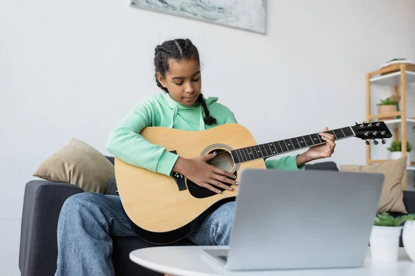 Concentrated african american teenage girl learning to play guitar near blurred laptop — Stock Photo