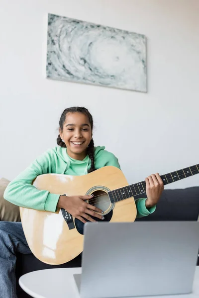 Cheerful african american teen girl with acoustic guitar looking at camera near blurred laptop — Stock Photo