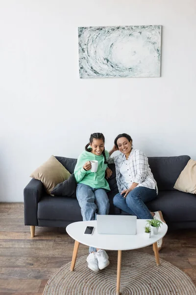 Lächelnde afrikanisch-amerikanische Mädchen mit einer Tasse Tee, die gemeinsam mit Mama einen Film auf dem Laptop anschaut — Stockfoto