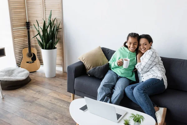 Happy african american girl holding cup of tea while watching movie on laptop near mother — Stock Photo