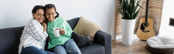 Afrikanisch-amerikanische Teenager-Mädchen mit Tasse Tee umarmt mit glücklicher Mutter auf der Couch zu Hause, Banner — Stockfoto