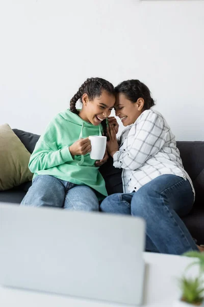 Excited african american girl with cup of tea laughing together with mom while watching movie on blurred laptop — Stock Photo