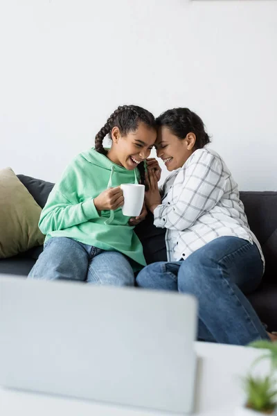 Excitada chica afroamericana con taza de té riendo junto con mamá mientras ve la película en un ordenador portátil borroso - foto de stock