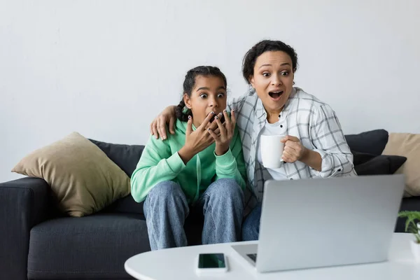 Femme afro-américaine choquée avec fille adolescente regardant un film sur ordinateur portable flou — Photo de stock