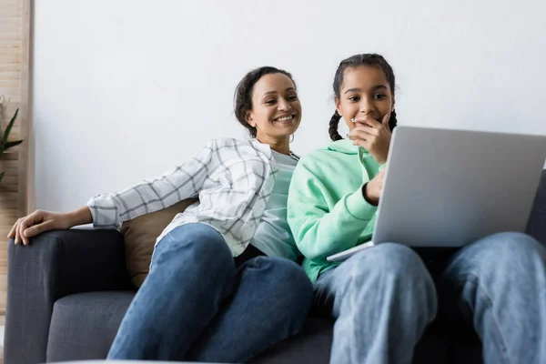 Erstaunt afrikanisch-amerikanische Teenager-Mädchen, die Mund mit der Hand bedecken, während sie Film auf Laptop in der Nähe lächelnde Mutter ansehen — Stockfoto