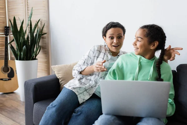 Étonnante femme afro-américaine pointant du doigt un ordinateur portable tout en regardant un film avec une adolescente joyeuse — Photo de stock