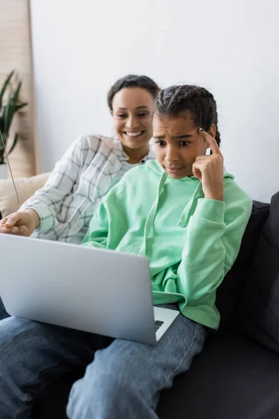 Reflexiva afroamericana chica viendo película en el ordenador portátil cerca sonriente madre - foto de stock