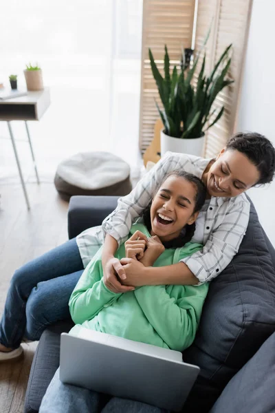 Feliz africana americana mujer abrazando riendo hija mientras viendo película en portátil en casa - foto de stock