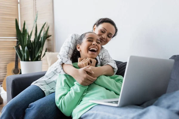 African american woman hugging laughing daughter while watching film on laptop — Stock Photo