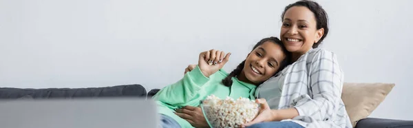 Alegre afroamericana mujer y adolescente chica viendo película en portátil cerca de tazón de palomitas de maíz, bandera - foto de stock