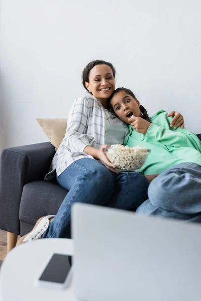 Sonriente mujer afroamericana con palomitas de maíz viendo la película en un ordenador portátil borroso con hija sorprendida - foto de stock