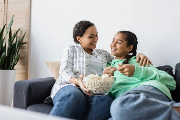 Mujer afroamericana feliz con tazón de palomitas de maíz sentado en el sofá con hija adolescente - foto de stock