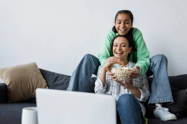 Happy african american woman with teenage daughter watching movie on laptop near bowl of popcorn — Stock Photo