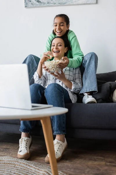 Cheerful african american mother and daughter eating popcorn while watching film on blurred laptop — Stock Photo