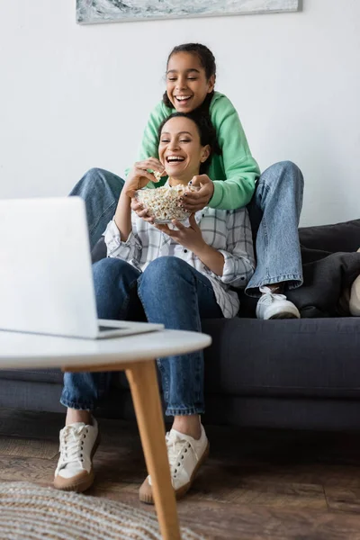 Fröhliche afrikanisch-amerikanische Mutter und Tochter essen Popcorn, während sie Film auf verschwommenem Laptop ansehen — Stockfoto