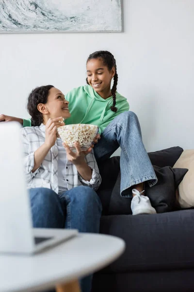 Fröhliches afrikanisch-amerikanisches Mädchen schaut Mutter mit Schüssel Popcorn in der Nähe von verschwommenem Laptop an — Stockfoto