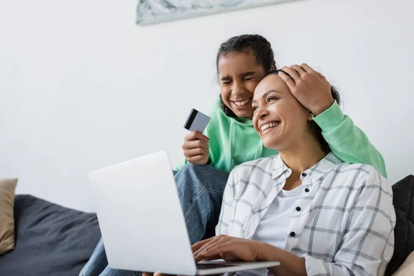 Excited african american teen girl with closed eyes girl holding credit card near smiling mom and laptop — Stock Photo