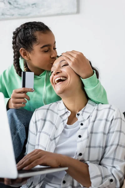 Afrikanerin küsst fröhliche Mutter auf die Stirn, während sie Kreditkarte in der Nähe eines verschwommenen Laptops hält — Stockfoto