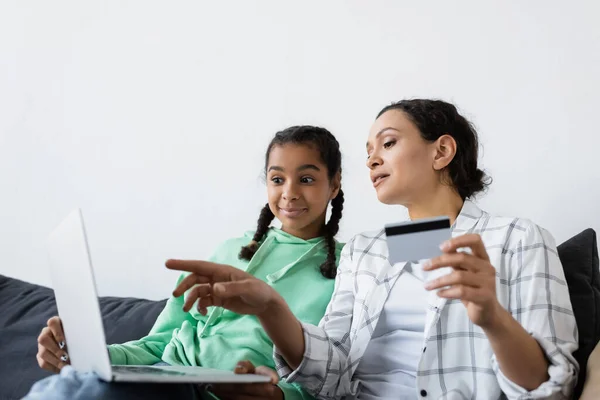 Mujer afroamericana sosteniendo tarjeta de crédito y señalando con el dedo a la computadora portátil cerca de hija sorprendida - foto de stock