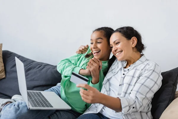 Sonriente afroamericana mujer sosteniendo tarjeta de crédito cerca de la computadora portátil y emocionada hija - foto de stock