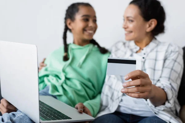 Blurred african american woman holding credit card near laptop and happy teenage daughter — Stock Photo