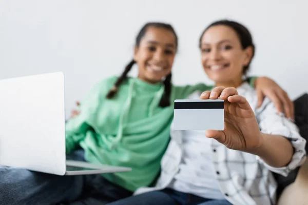 Selective focus of credit card in hand of blurred african american woman sitting near laptop and teen daughter — Stock Photo