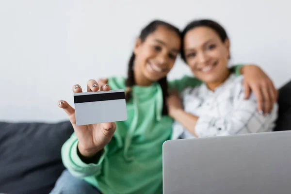 Розмита афроамериканська дівчина-підліток тримає кредитну картку, обіймаючи маму біля ноутбука — стокове фото