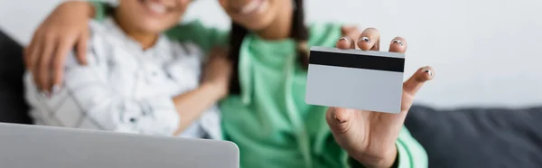 Cropped view of blurred african american girl holding credit card near laptop and mom, banner — Stock Photo