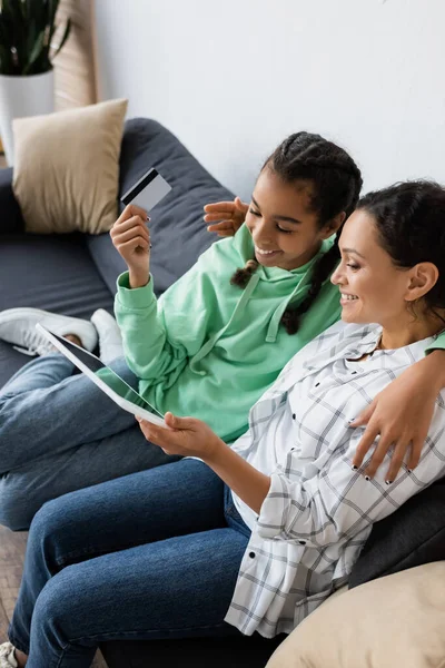 Happy african american girl holding credit card near mom with digital tablet — Stock Photo