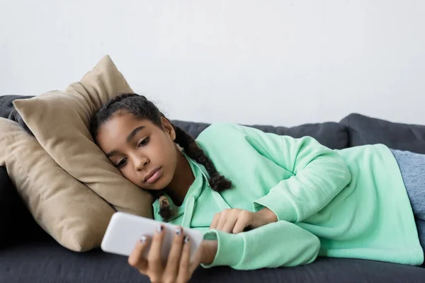 Upset african american teen girl using mobile phone while lying on sofa — Stock Photo