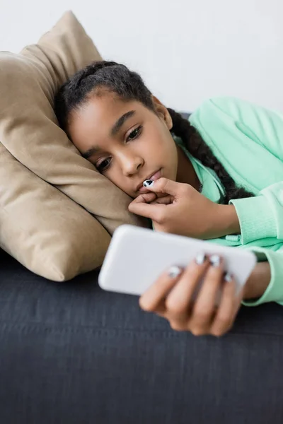 Afrikanisch-amerikanisches Teenager-Mädchen schaut auf verschwommenes Smartphone, während es zu Hause auf der Couch liegt — Stockfoto