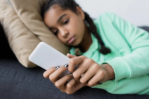 Blurred african american teen girl in bad mood messaging on mobile phone while lying on couch — Stock Photo