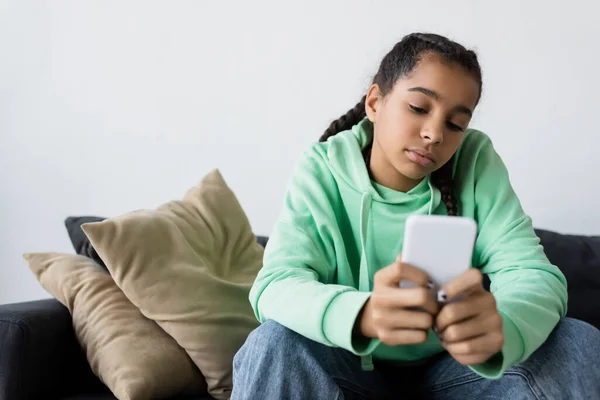 Frustrado afroamericano adolescente chica mensajería en el teléfono móvil borroso mientras está sentado en el sofá - foto de stock
