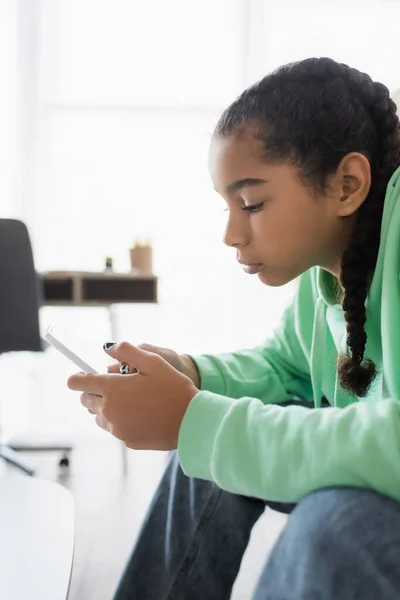 Vista laterale della triste ragazza adolescente africana americana che chatta sul telefono cellulare a casa — Foto stock